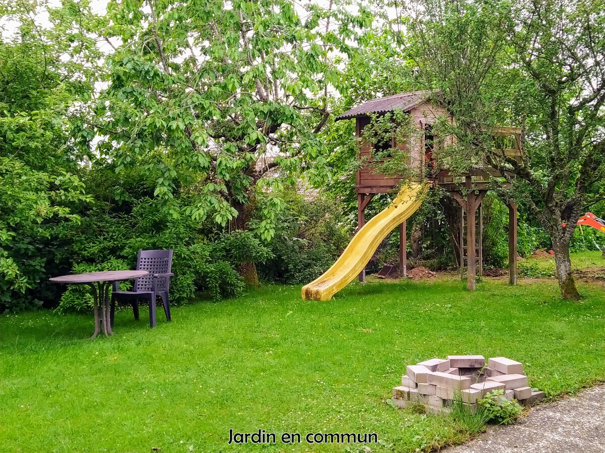 A shared garden area is depicted, featuring a play structure with a yellow slide under a canopy of lush green trees. A small table and chairs are situated on the grassy lawn, with a stone fire pit arranged nearby.
