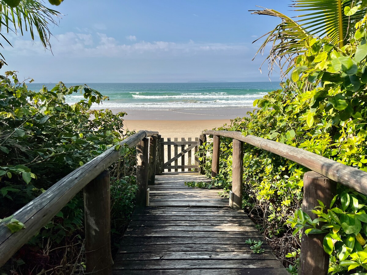 A wooden pathway leads to a sandy beach, flanked by lush greenery on either side. Gentle waves can be seen in the distance under a clear blue sky, with a wooden gate at the end of the pathway providing access to the shore.