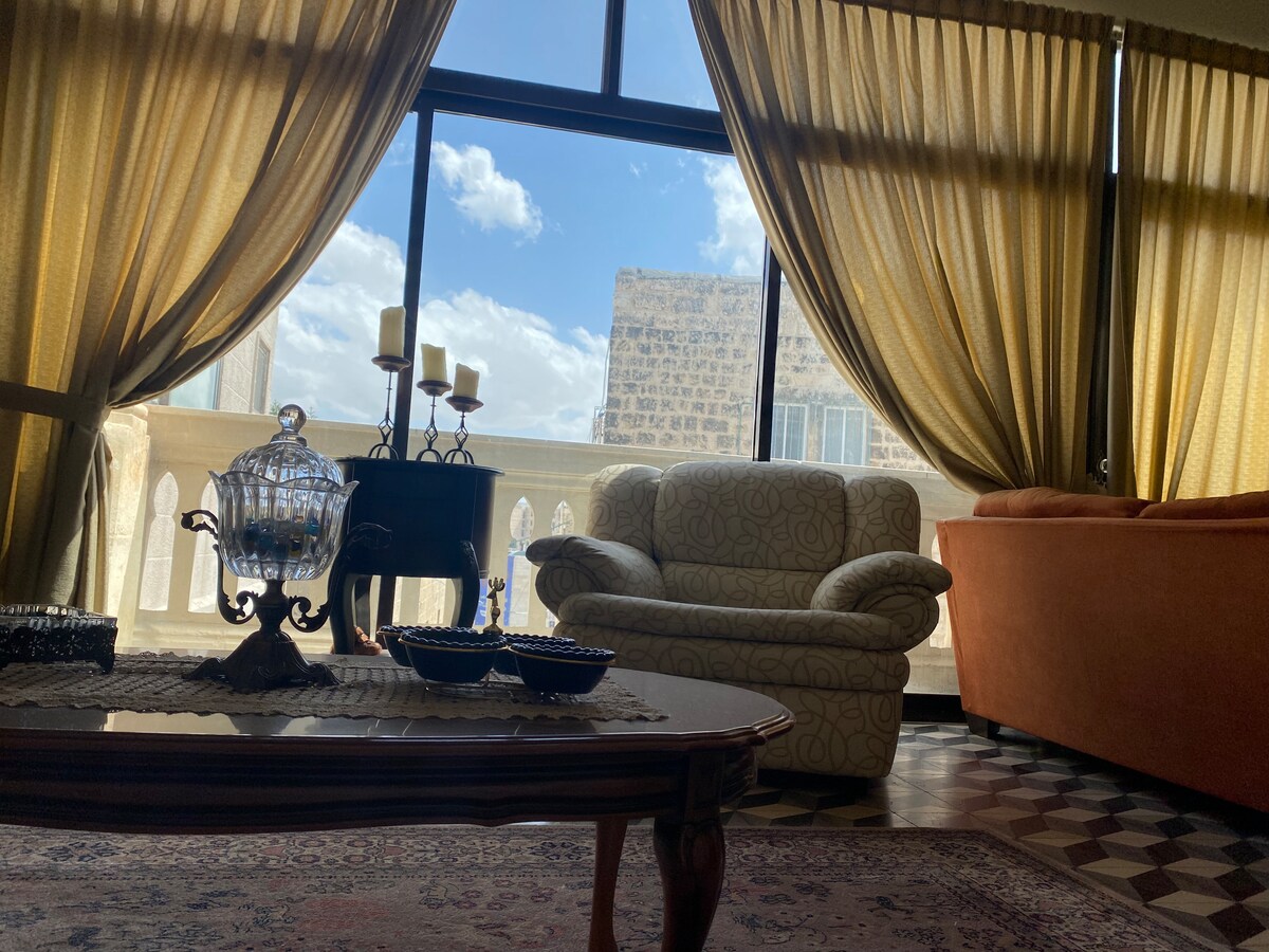 A spacious living area is illuminated by natural light streaming through large windows. Soft curtains frame the view, while a cozy couch and patterned armchair provide comfortable seating. A wooden coffee table with decorative items complements the intricate floor design.