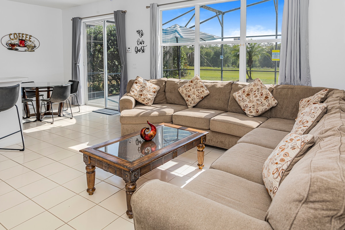A spacious living area is illuminated by natural light streaming through large glass doors, revealing views of the outdoors. A large, comfortable sectional sofa adorned with decorative pillows surrounds a wooden coffee table. A dining table with modern chairs is positioned nearby.