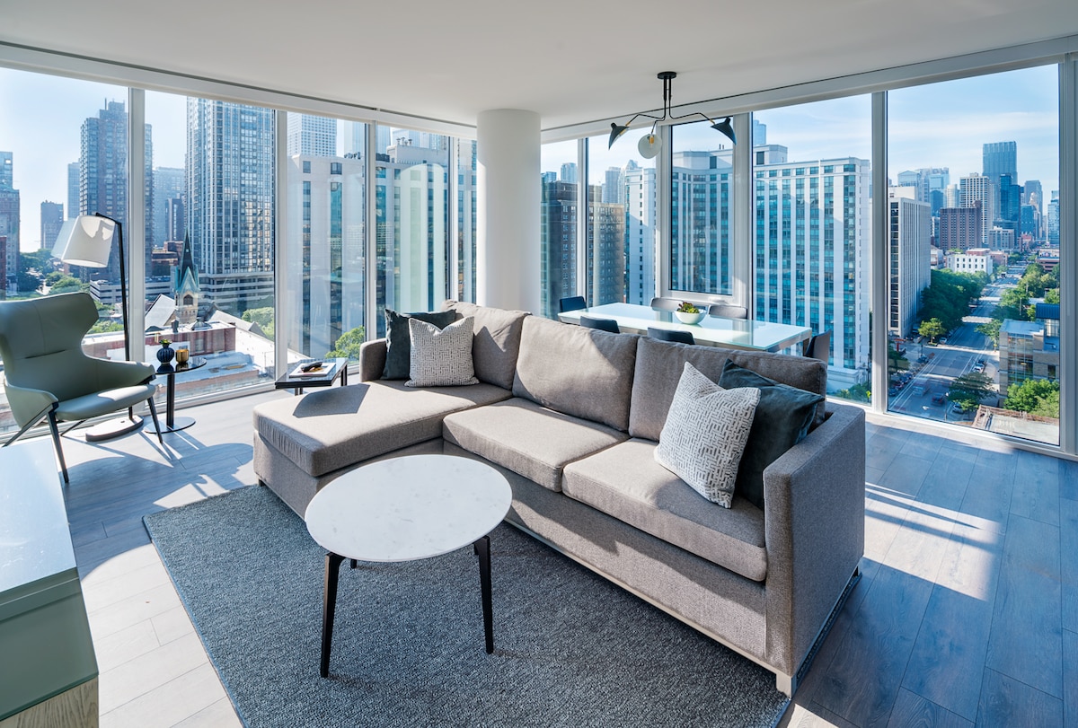 A spacious living area is seen with floor-to-ceiling windows offering city views. A light gray sectional sofa sits on a soft area rug, accompanied by a round coffee table. Natural light fills the room, highlighting the modern décor and open layout.