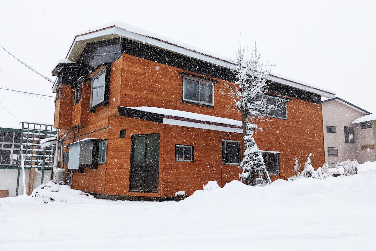 The exterior of a wooden chalet is surrounded by a blanket of fresh snow. The building features multiple windows, and a large entrance area is visible. Snowflakes fall gently, creating a serene winter atmosphere. Trees are lightly dusted with snow, complementing the cozy appearance.