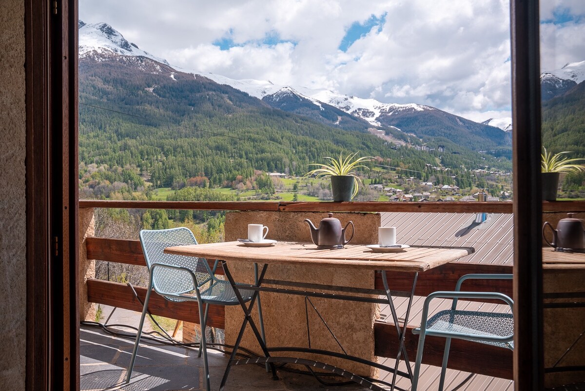 A spacious balcony is visible, featuring a table set with two cups and a teapot. Two chairs are placed beside the table, offering an inviting spot to enjoy the surrounding mountain views and natural landscape.