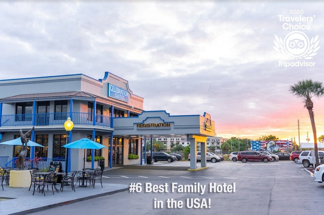 A welcoming hotel facade is presented with a registration area marked by a bright blue awning. Outdoor seating is arranged at tables, with palm trees positioned nearby. The sky exhibits soft pastel colors during sunset, creating a calm ambiance around the property.