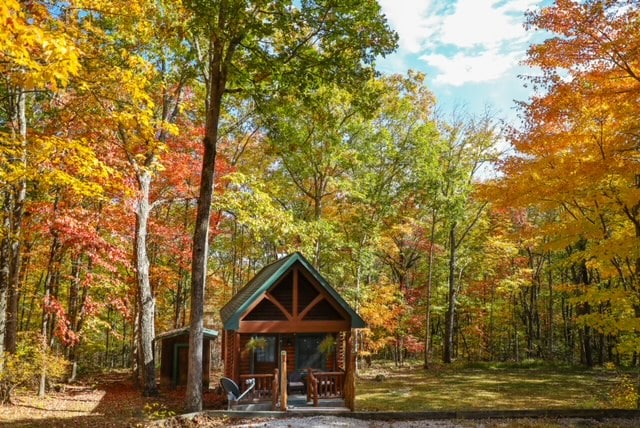 The log cabin features a peaked roof and large windows that invite natural light. Surrounded by autumn foliage, vibrant reds and oranges create a picturesque setting. A small porch with two chairs adds to the inviting outdoor space, enhancing the cabin's secluded charm.