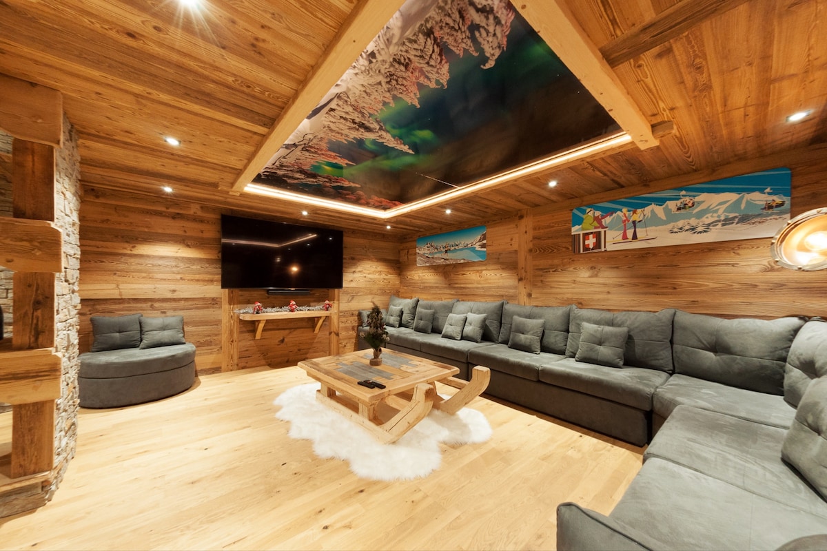 A spacious living area features a large L-shaped gray sectional sofa surrounding a central wooden coffee table. The wood-paneled walls and floor add warmth, while a striking mountain-themed ceiling mural enhances the ambiance. A flat-screen TV is mounted for entertainment.