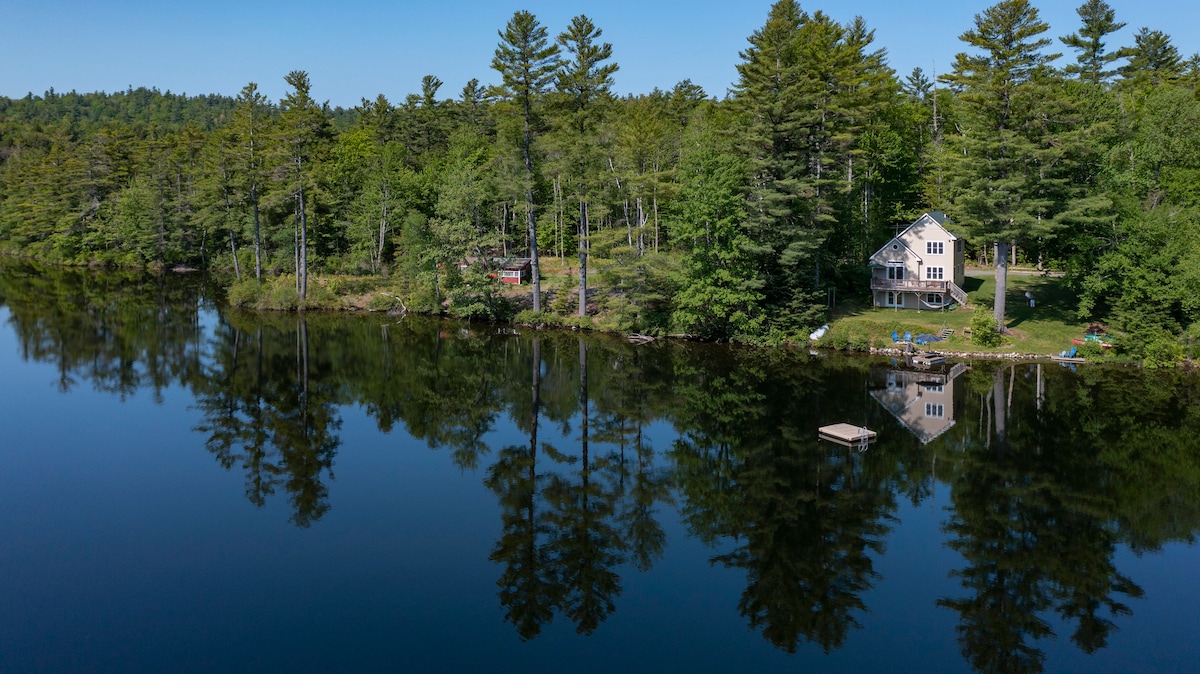 A serene lakefront cottage is depicted, surrounded by lush greenery and tall trees. The calm lake mirrors the landscape, enhancing the tranquil atmosphere. A small dock extends into the water, inviting relaxation. The property showcases a private retreat, perfect for peaceful family gatherings.