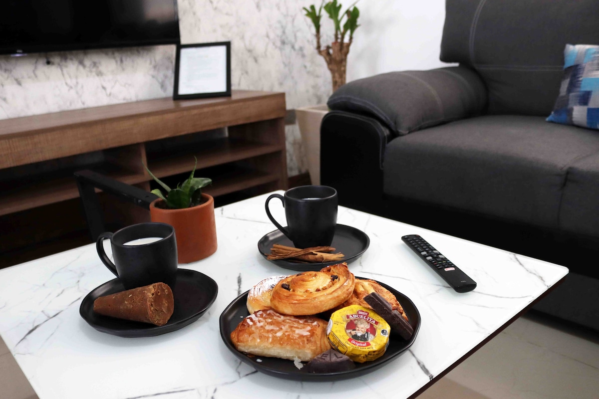 A stylish coffee table displays a round plate with assorted baked goods, including a pastry and a chocolate treat. Two black cups are placed on the table, accompanied by a small potted plant. In the background, a comfortable gray sofa and a television are visible.