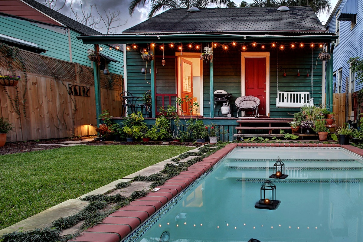 A cozy outdoor space features a cocktail pool with a sun shelf, lined with decorative lanterns. The green cottage is framed by well-maintained gardens and a wooden fence, with string lights illuminating the entrance and porch area, creating a welcoming atmosphere.
