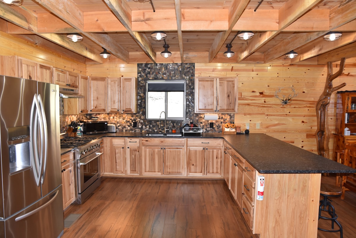 A spacious kitchen features natural wood cabinetry and a large island with dark countertops. Stainless steel appliances are arranged for convenience, and a decorative stone backsplash adds visual interest. A window allows natural light to illuminate the space.