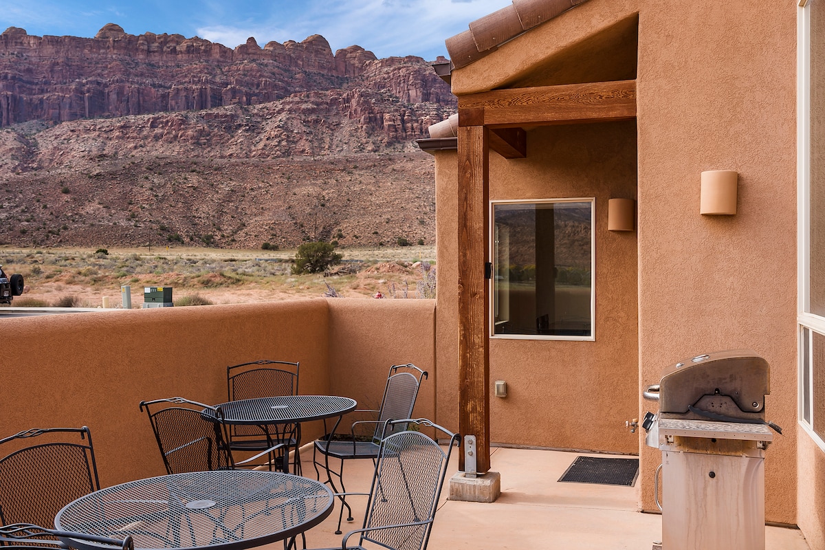 A private patio is displayed with multiple outdoor tables and chairs, offering a space for relaxation. A grill is positioned nearby, and a large window allows for views of the surrounding rugged red rock landscape.