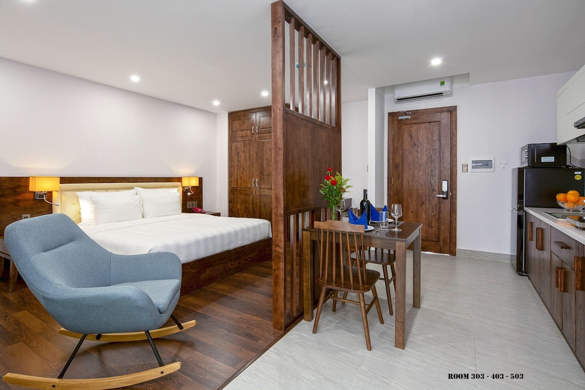 A spacious room is divided by a wooden partition, featuring a comfortable bed and a dining area with a wooden table and chairs. A blue rocking chair is positioned nearby. A kitchenette and a small vase with flowers are visible, contributing to a welcoming ambiance.