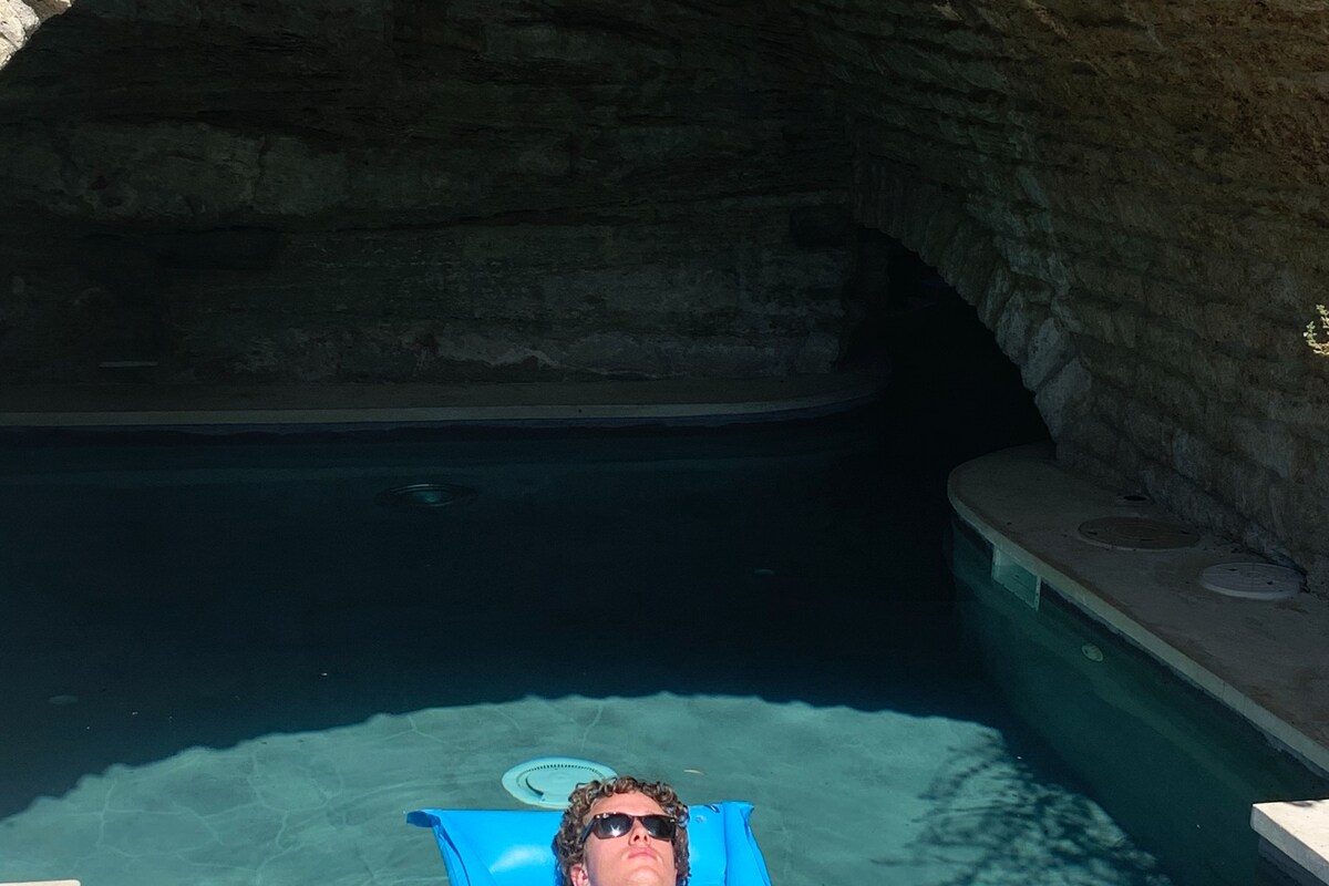 A grotto pool is surrounded by natural stone walls, providing a serene ambiance. The water appears calm and inviting, reflecting the contours of the arching ceiling above. A floating lounge is positioned on the surface, enhancing the relaxation potential of the space.