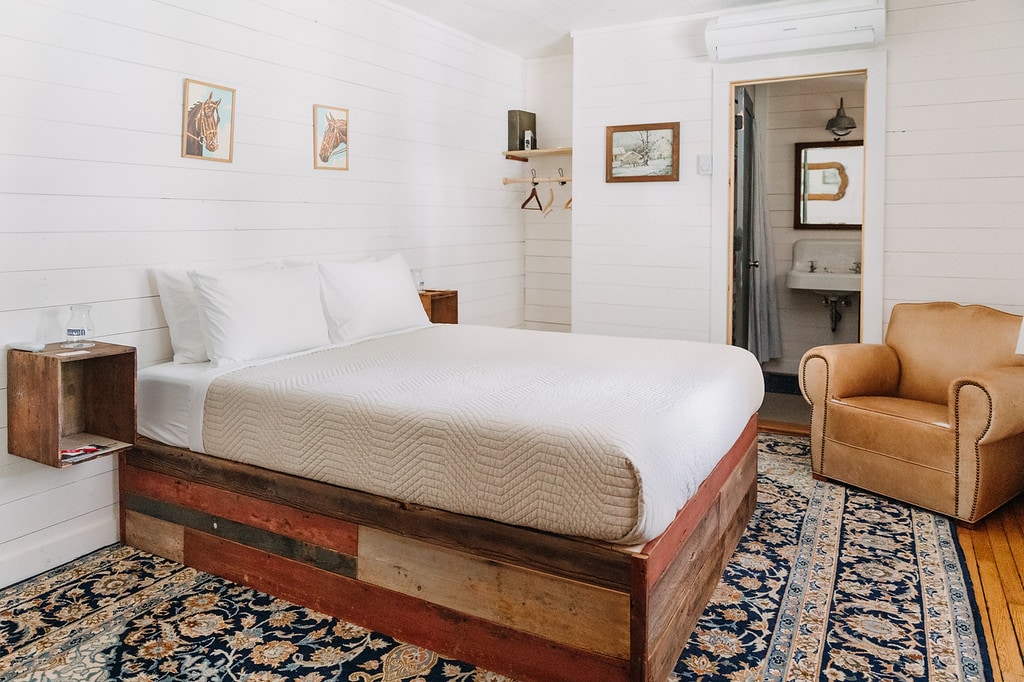 A spacious sleeping area is presented, featuring a large bed with a neutral-colored quilt and upholstered headboard. An armchair is positioned near a wooden nightstand, and artwork adorns the walls. A soft, patterned rug complements the wood accents in the room.