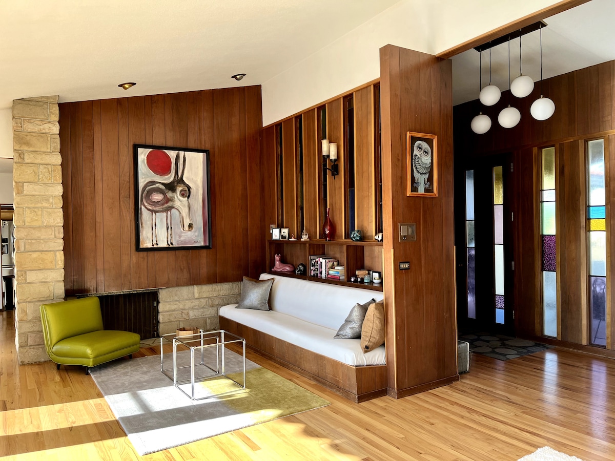 A mid-century modern living area features warm wood paneling and a vibrant piece of artwork. A sleek white bench is positioned along the wall, complemented by a small coffee table and a bright green armchair. Colorful stained glass accents are visible near the entrance.