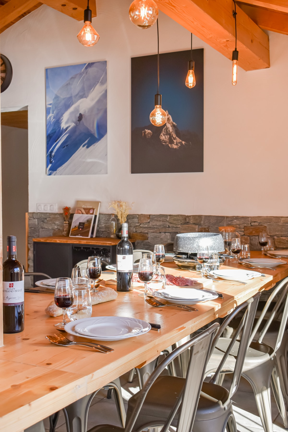 A spacious dining area features a long wooden table set for a meal, complete with plates and glasses. Above, pendant lights provide illumination, while artwork depicting mountain landscapes adorns the walls. The natural stone accents add warmth to the inviting space.