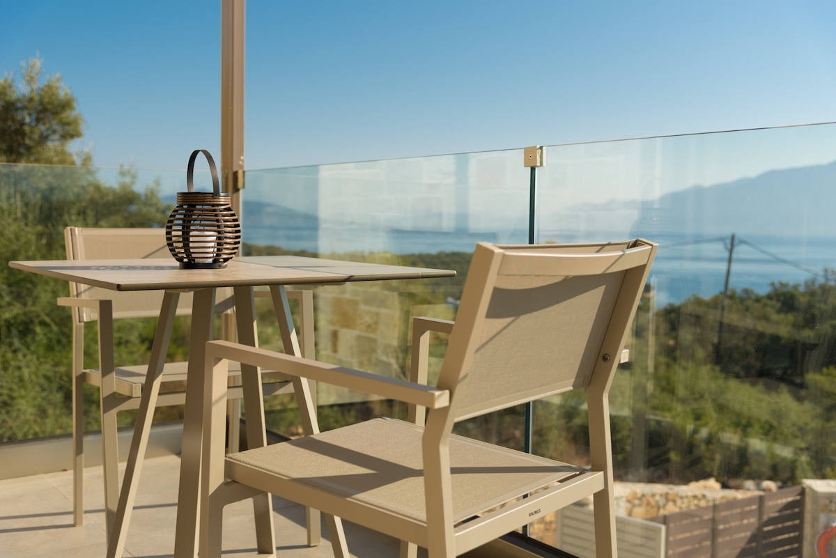 A cozy outdoor seating area is featured on a balcony, showcasing a small round table with two light-colored chairs. A woven lantern sits on the table, while a clear glass railing provides an unobstructed view of the distant landscape and water.