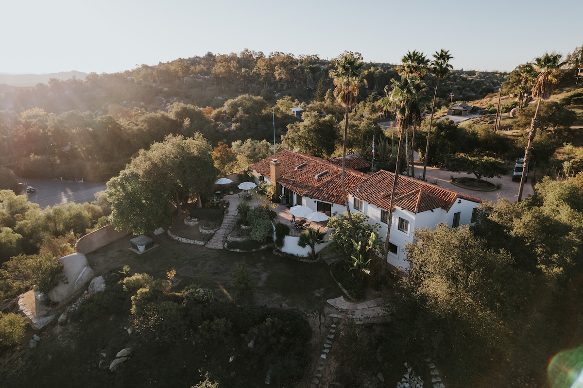 An aerial view captures the expansive layout of the property, showcasing the main house surrounded by lush greenery and palm trees. The vibrant red-tiled roof contrasts with the natural landscape, while outdoor seating areas can be seen nestled among the gardens.