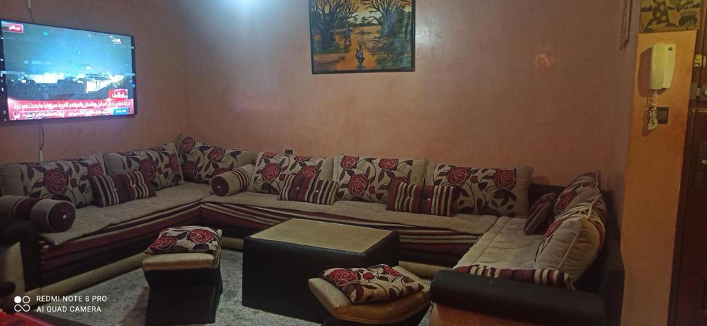 A spacious living area features a large sectional sofa adorned with floral patterns. A central coffee table is surrounded by smaller seats. A flat-screen TV is mounted on the wall, and a decorative painting hangs above the sofa, contributing to the room's inviting ambiance.