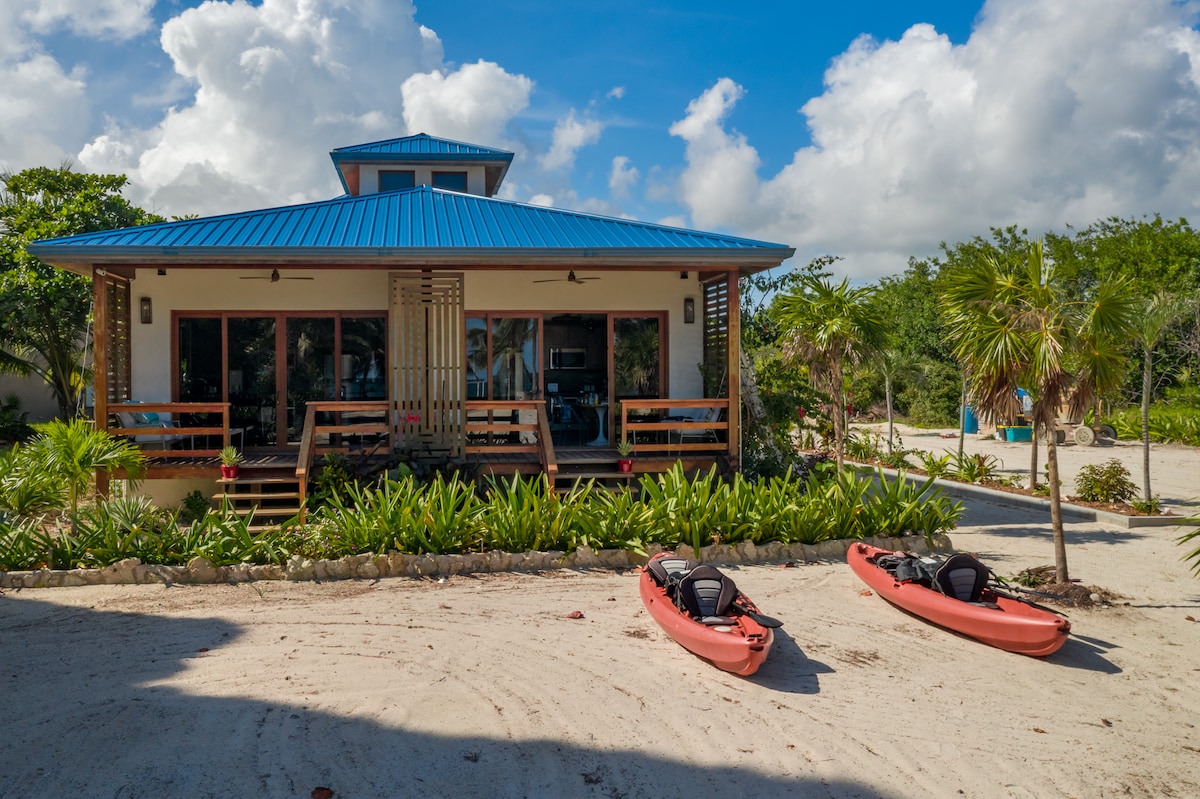 A charming beachfront villa is framed by lush tropical landscaping and sandy pathways. Two kayaks are positioned on the sandy area in front, ready for adventure. The villa's blue roof contrasts beautifully with the white clouds above, creating a relaxed and inviting atmosphere.