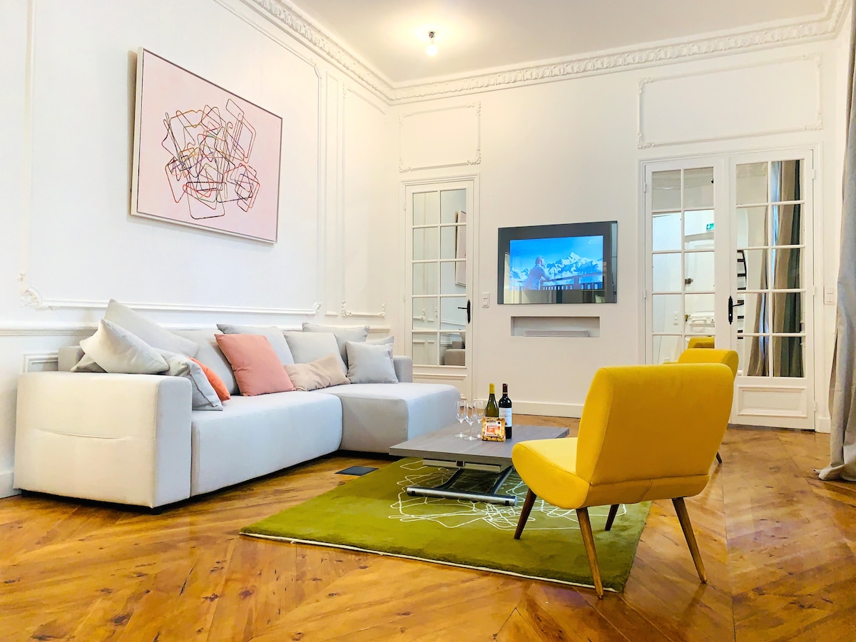 A spacious living area features a comfortable gray sofa adorned with pastel cushions. A bright yellow chair complements a low coffee table placed on a green rug. A large wall-mounted TV is visible, and elegant glass doors open to another room, allowing natural light to fill the space.