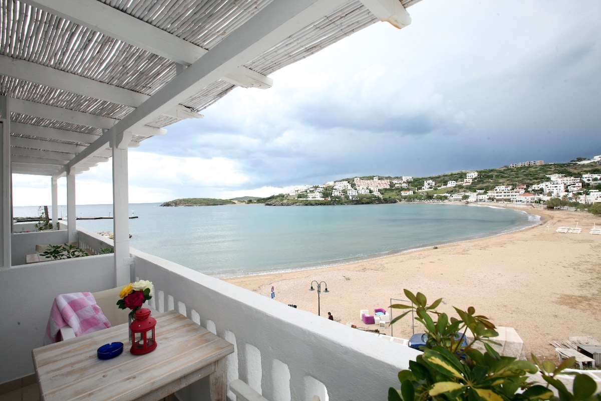 A serene balcony offers views of the sandy beach and calm waters, framed by coastal hills and seaside buildings. A small wooden table with a flower vase and a lantern adds a welcoming touch, while overhead wooden beams provide shade.