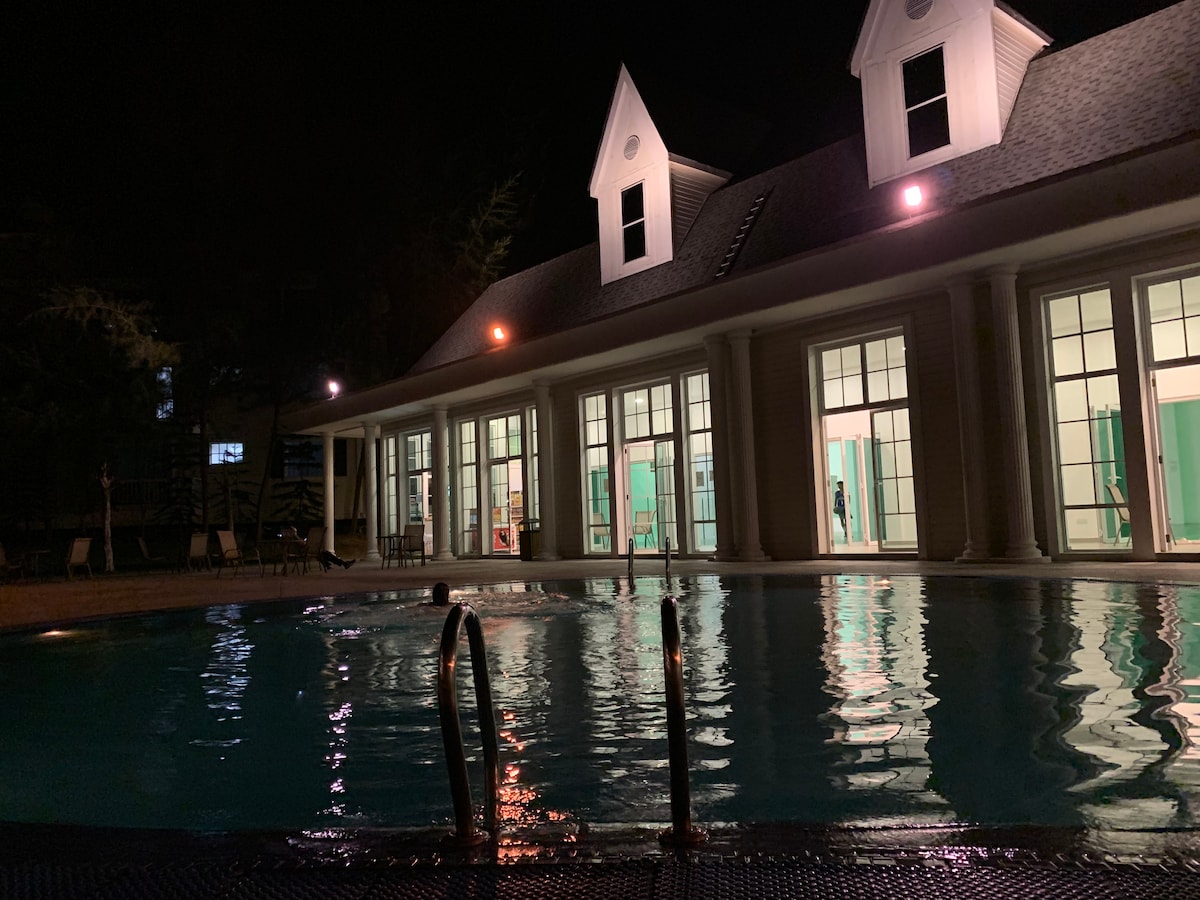 An outdoor swimming pool is illuminated at night, reflecting light from surrounding buildings. The pool area features steps leading into the water, with large glass doors on one side revealing a spacious indoor area with green accents.