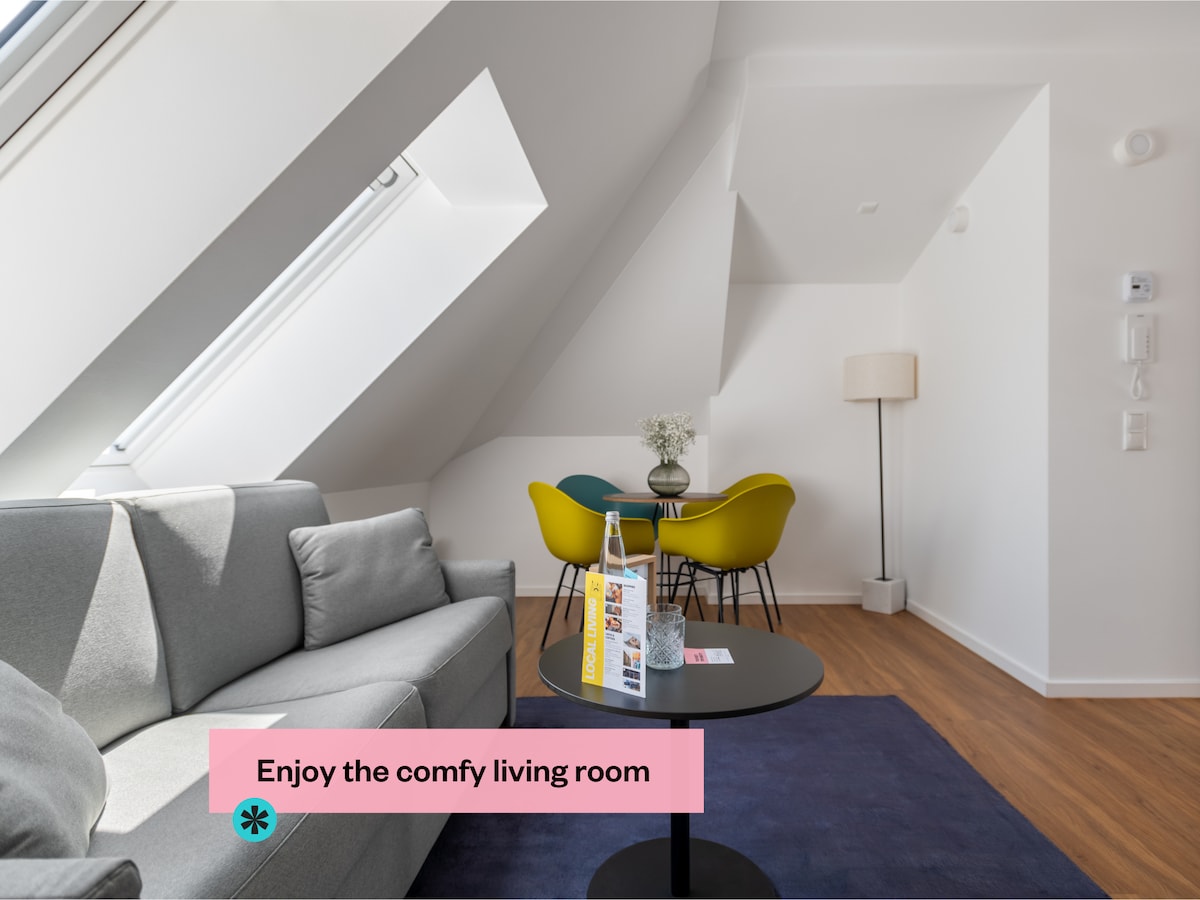 The living room features a gray sofa complemented by a dark coffee table. A small dining area is visible with colorful chairs and a lamp, positioned below sloped ceilings. Natural light enters through large windows, highlighting the modern decor and warm wooden flooring.