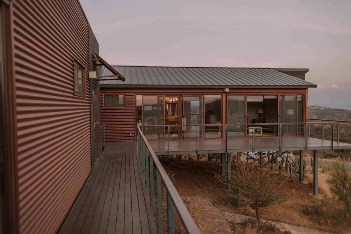 An exterior view of the villa showcases a modern structure with a sloped metallic roof and a maroon corrugated facade. A wooden deck offers access to expansive glass doors, revealing the interior space. The surrounding landscape features sparse vegetation in a natural setting.
