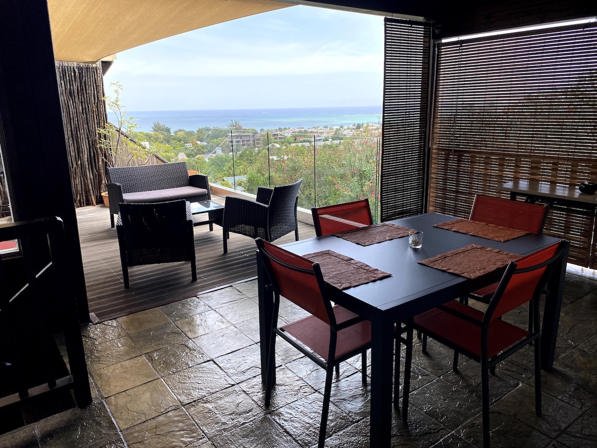 An outdoor dining area is presented, featuring a table with four red chairs and placemats. A seating arrangement with two chairs and a small table is visible, along with a backdrop of lush greenery and a glimpse of the ocean. Natural light filters through shaded coverings.