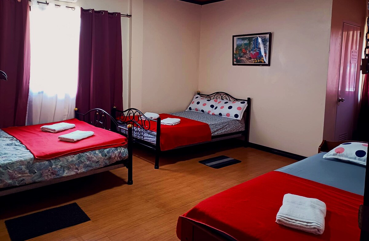 A room features three beds with bright red bed covers and patterned sheets. Soft towels are neatly placed on each bed. Window drapes filter natural light, illuminating the space without overwhelming it. A decorative wall picture adds a touch of color.