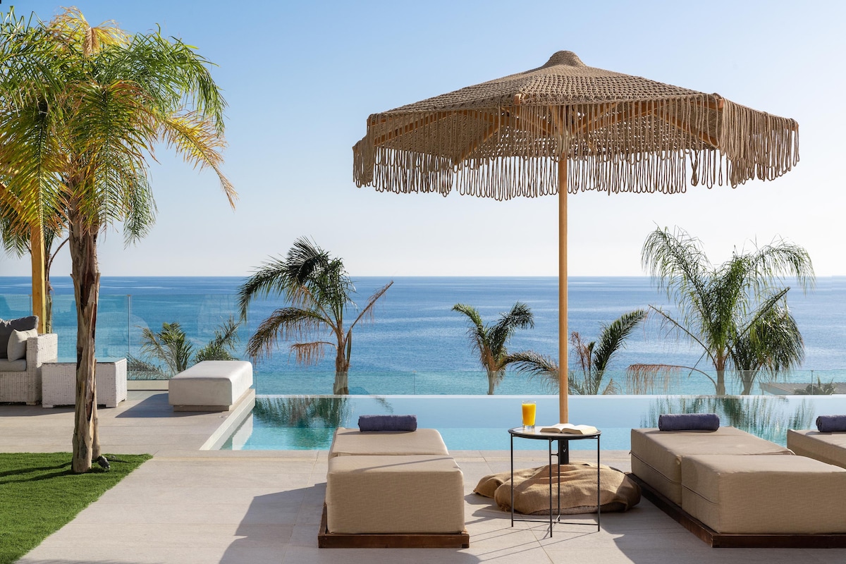 An outdoor seating area features sun loungers with soft cushions, accompanied by a round table. A large, fringed umbrella provides shade. The backdrop consists of an infinity pool that merges with expansive views of the tranquil sea and surrounding palm trees.