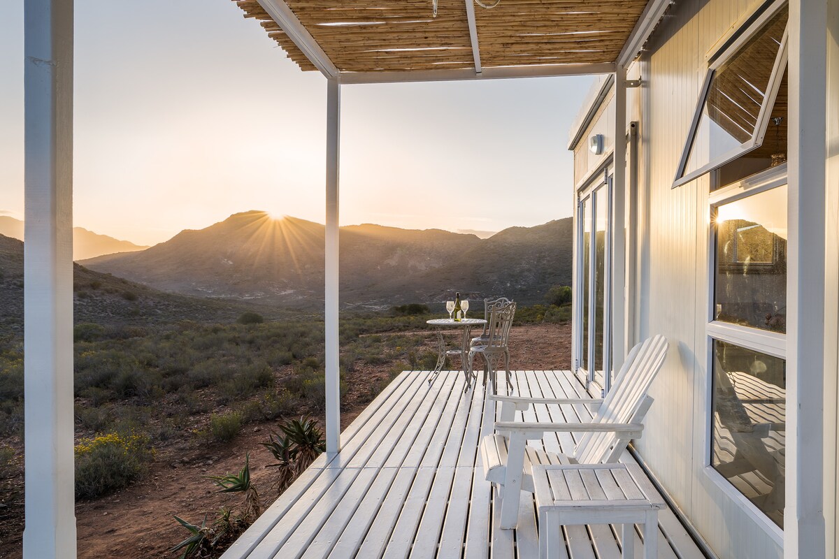 A spacious deck equipped with two white lounge chairs faces a breathtaking mountain view at sunset. A small table set with two glasses adds a touch of elegance, surrounded by unspoiled nature and gentle slopes.