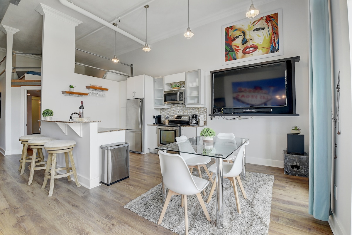 The open-concept living area features a glass dining table surrounded by white chairs, set on a plush rug. Modern kitchen appliances are visible, including a refrigerator and microwave. Exposed ceilings and large artwork contribute to the overall chic decor.