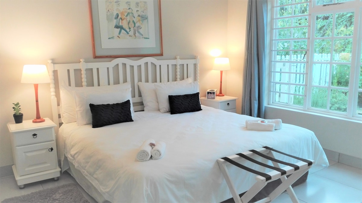 A well-appointed bedroom features a spacious bed adorned with crisp white linens. Two bedside lamps provide soft illumination. A decorative wall art piece hangs above the bed, and a plant accents a nearby nightstand. A window allows natural light to enter, framed by light curtains.