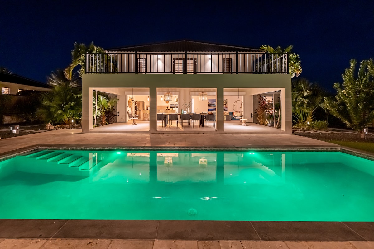 The villa is illuminated at night, featuring a spacious patio that overlooks a glowing turquoise pool. Lush greenery surrounds the property, enhancing its tropical setting. The second-floor balcony offers views of the main living area, which is visible through large glass doors.