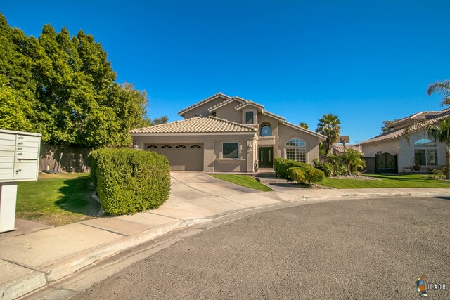 A spacious home with a light-colored exterior is set in a quiet neighborhood. The front yard features well-maintained shrubs and a manicured lawn. A driveway leads to a two-car garage, while the entrance is highlighted by large windows and a tiled roof under a clear blue sky.