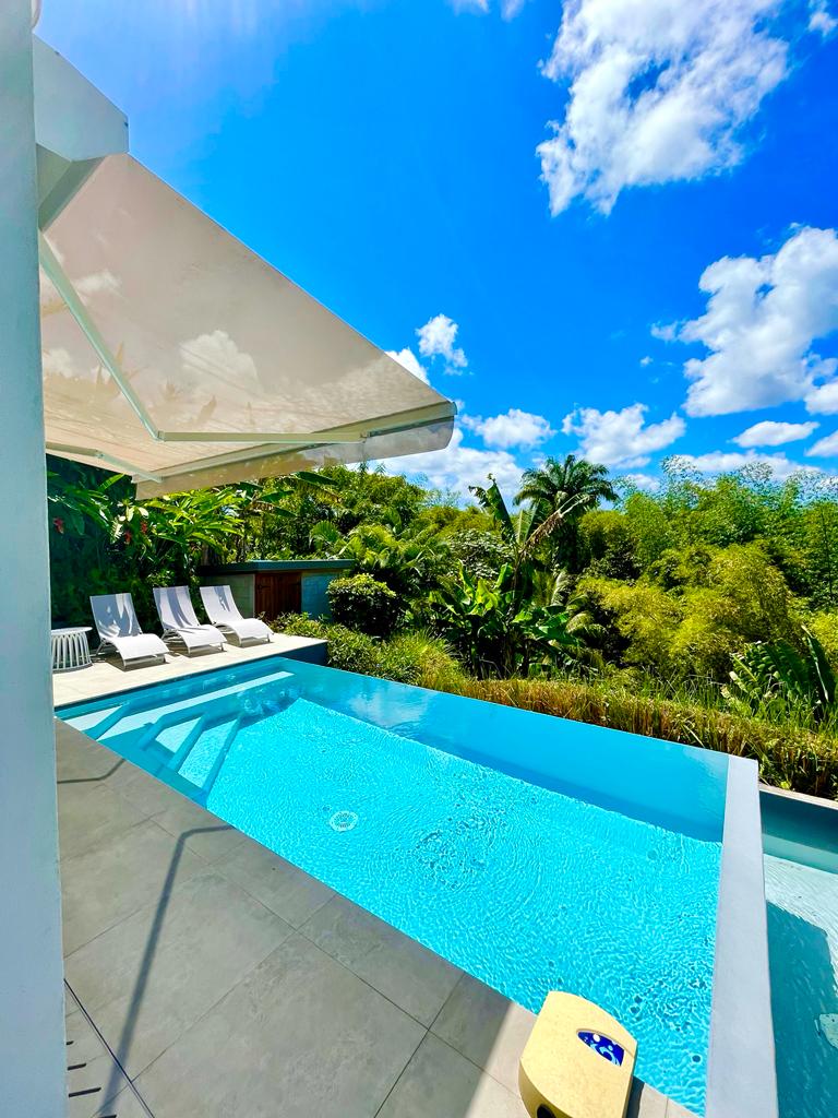A private swimming pool is featured with clear blue water, surrounded by tropical greenery. Sun loungers are positioned nearby, inviting relaxation. A white awning provides shade, and a bright sky with fluffy clouds is visible above, adding to the serene atmosphere.