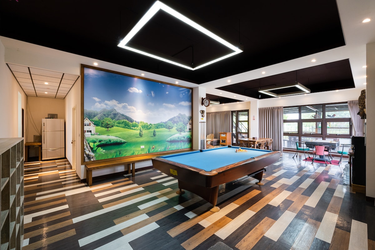A spacious recreation area features a billiards table prominently in the center. The backdrop displays a large mural of green hills and a serene landscape. Natural light filters through expansive windows, enhancing the inviting space decorated with a mix of wood and patterned flooring.