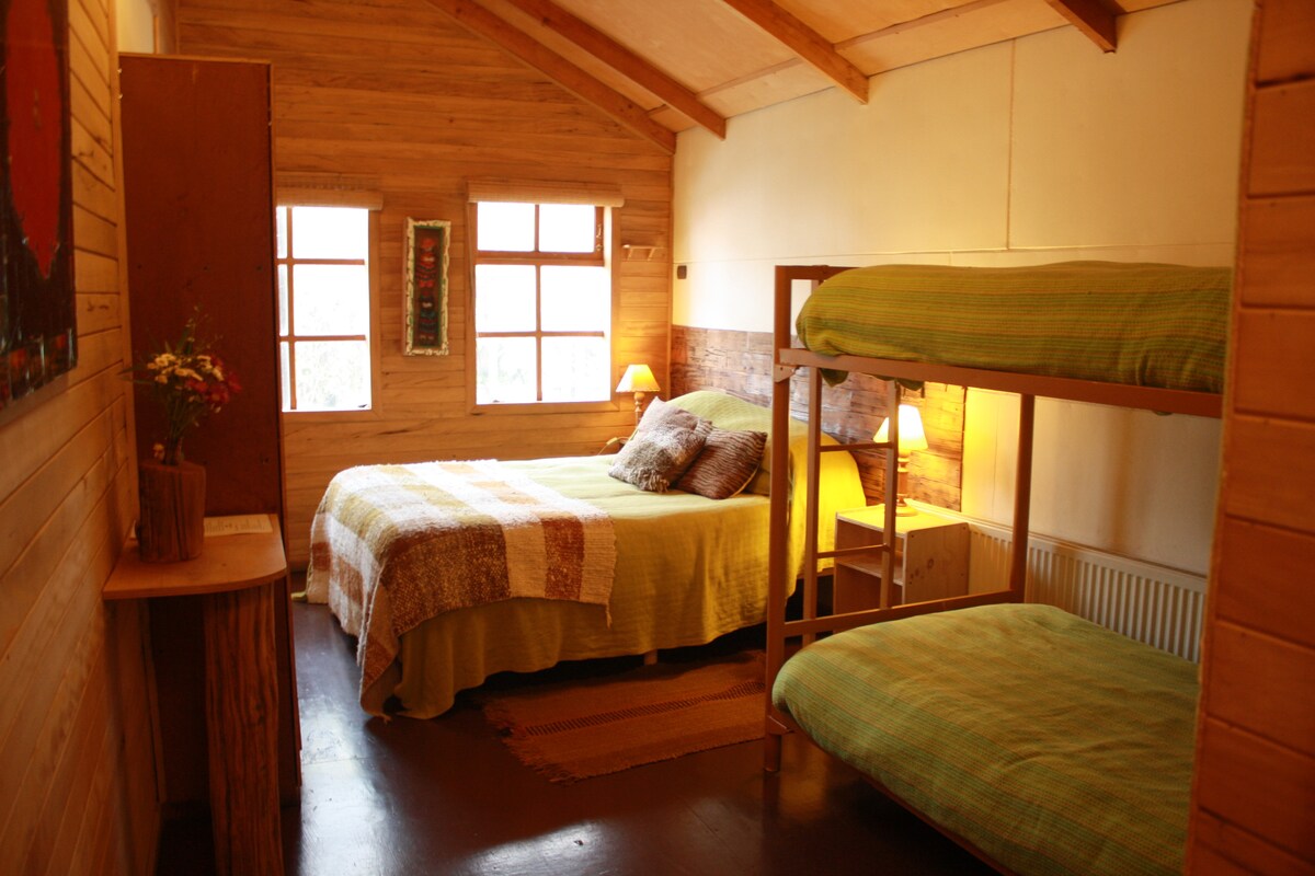 A family room features a double bed and a bunk bed, with natural wood paneling and large windows allowing natural light. The room includes two bedside lamps, a small wooden table, and a decorative wall hanging, creating a cozy atmosphere.