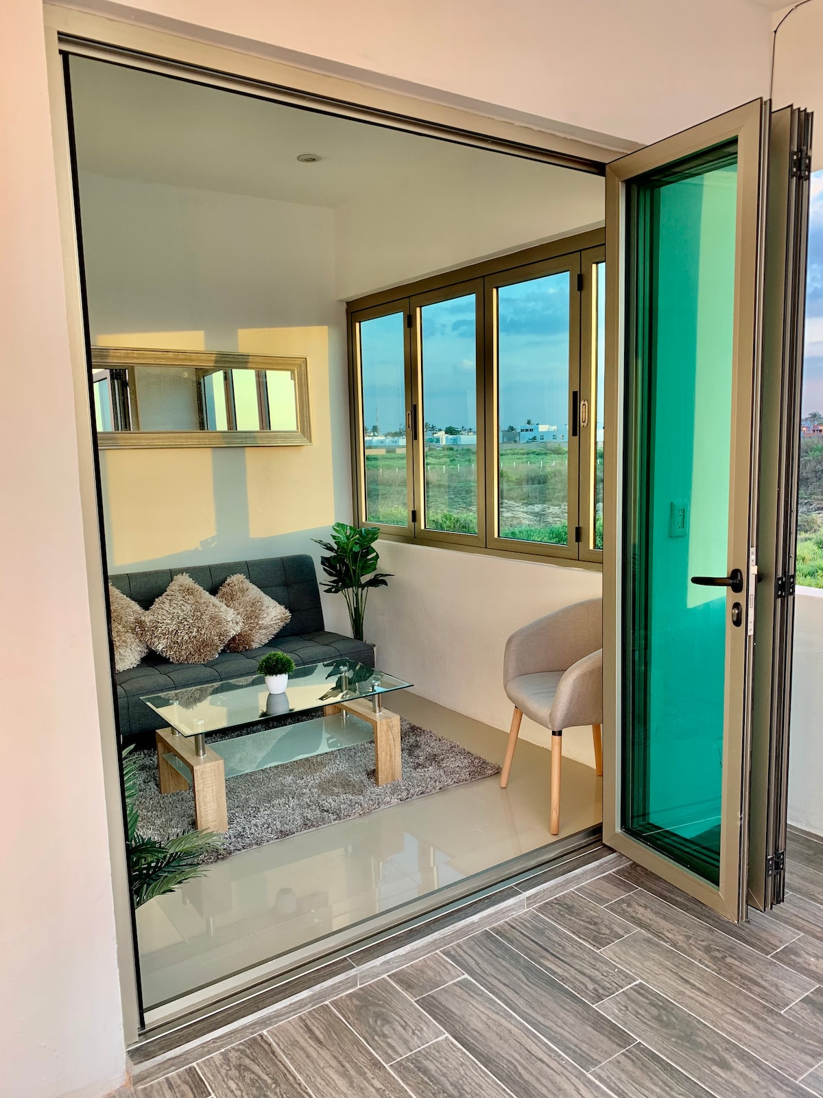 A small, bright space is displayed through a glass balcony door. A modern seating area features a gray couch with decorative cushions, complemented by a glass coffee table. Potted plants add greenery, while large windows offer views of the surrounding landscape.