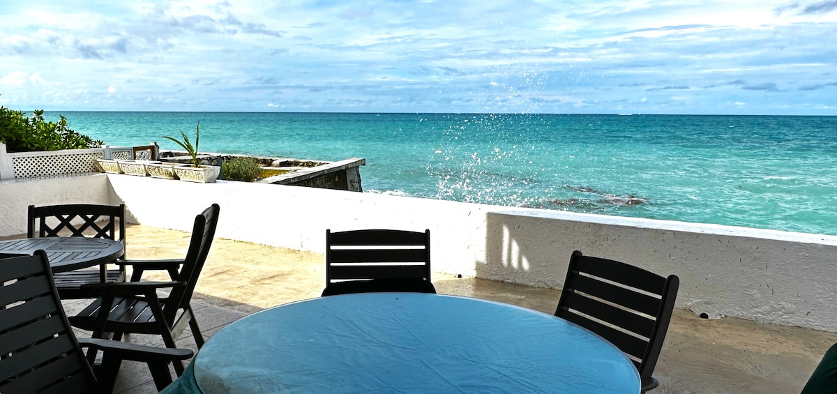 A seaside patio is shown, featuring a round table covered with a blue tablecloth. Black wooden chairs surround the table, with the ocean visible in the background. Waves gently splash against the rocky shore, creating a serene coastal view under a partly cloudy sky.