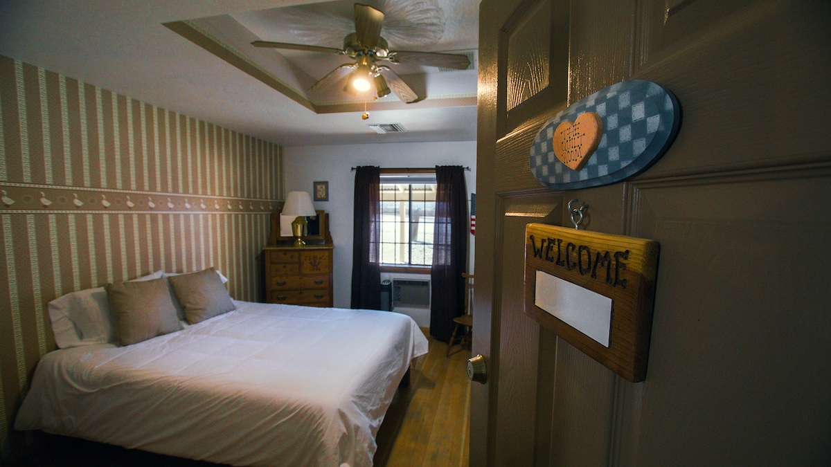 A private bedroom features a spacious bed dressed in crisp white linens. Against a backdrop of patterned wallpaper, a wooden dresser provides storage. Natural light enters through a window adorned with dark curtains. A welcome sign hangs on the door, inviting guests inside.