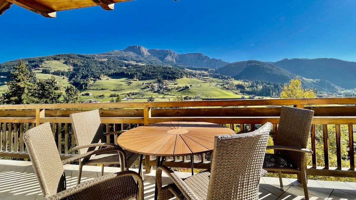 An outdoor terrace is featured with a round wooden table and five rattan chairs arranged around it. The panoramic view showcases rolling green hills and mountains under a clear blue sky.