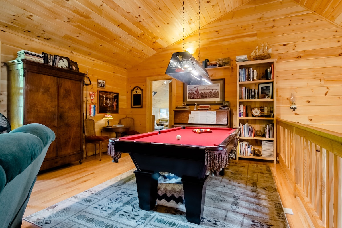 A cozy loft area features a red felt pool table beneath a stylish overhead light. Wooden walls and ceiling contribute to the rustic charm. An armoire and bookshelves are present, surrounded by inviting seating, creating a relaxed environment for leisure activities.