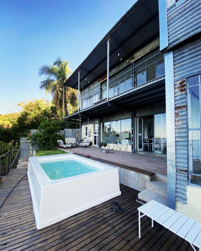 A modern building made of metal and glass sits amidst greenery. An inviting white plunge pool is situated on a wooden deck, complemented by lounge chairs and small dining tables. Large glass doors provide easy access to the indoor space, enhancing the connection with nature.