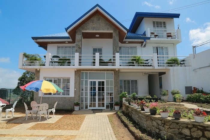 A multi-story building with a sloped blue roof is presented. A spacious terrace features colorful umbrellas over white tables and chairs. Surrounding flower beds and potted plants enhance the welcoming appearance. Large windows allow natural light into the interior space visible through the glass doors.
