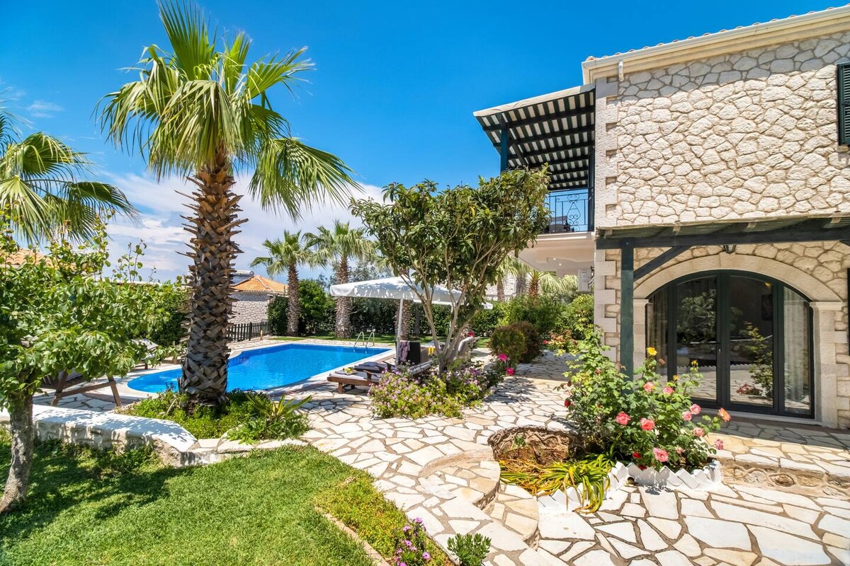 A stone villa is surrounded by a lush garden with palm trees and colorful flowers. A large swimming pool reflects the clear blue sky, with sun loungers positioned nearby. The outdoor area features a stone pathway leading to an inviting outdoor seating space under a pergola.