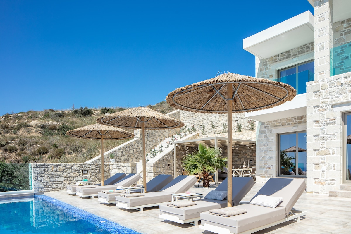 A sunlit terrace features luxury sunbeds beneath thatched umbrellas, providing shade next to the private infinity swimming pool. The backdrop consists of natural stone walls and a landscaped area with greenery, complemented by clear blue skies.