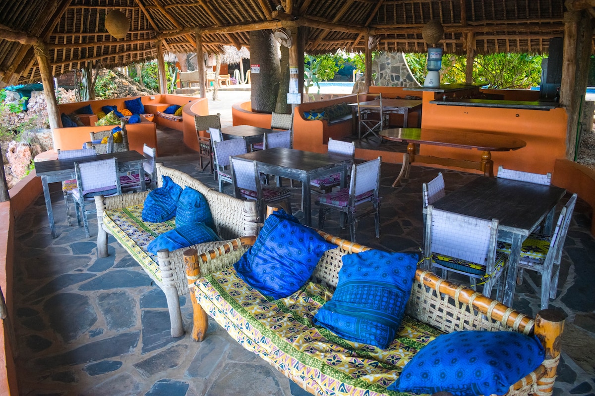An open dining area features numerous wooden tables and seating arranged under a thatched roof. Colorful cushions rest on a woven bench, complemented by vibrant textiles. Natural stone flooring adds to the rustic charm, creating a comfortable gathering space that blends with the surrounding environment.